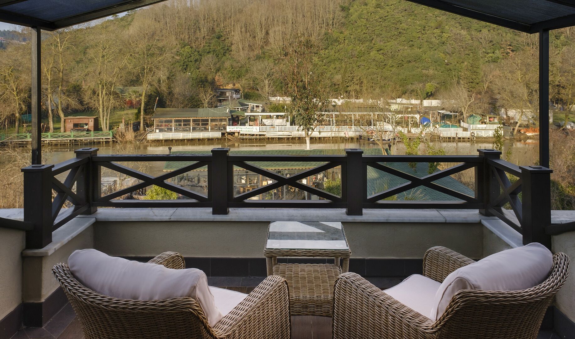 Un área de estar al aire libre cubierta con mobiliario de mimbre y vistas a un río con barcos y edificios al fondo.