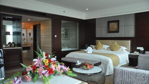 A hotel room with a large bed, a desk with a computer, a chair, a small round table with a fruit bowl, and a floral arrangement. - Conifer Garden Hotel (Haikou)