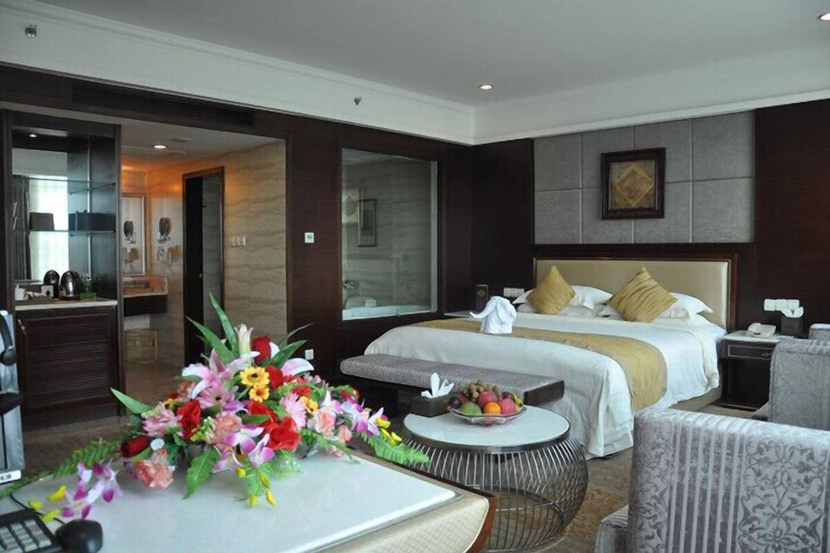 A hotel room with a large bed, a desk with a computer, a chair, a small round table with a fruit bowl, and a floral arrangement.