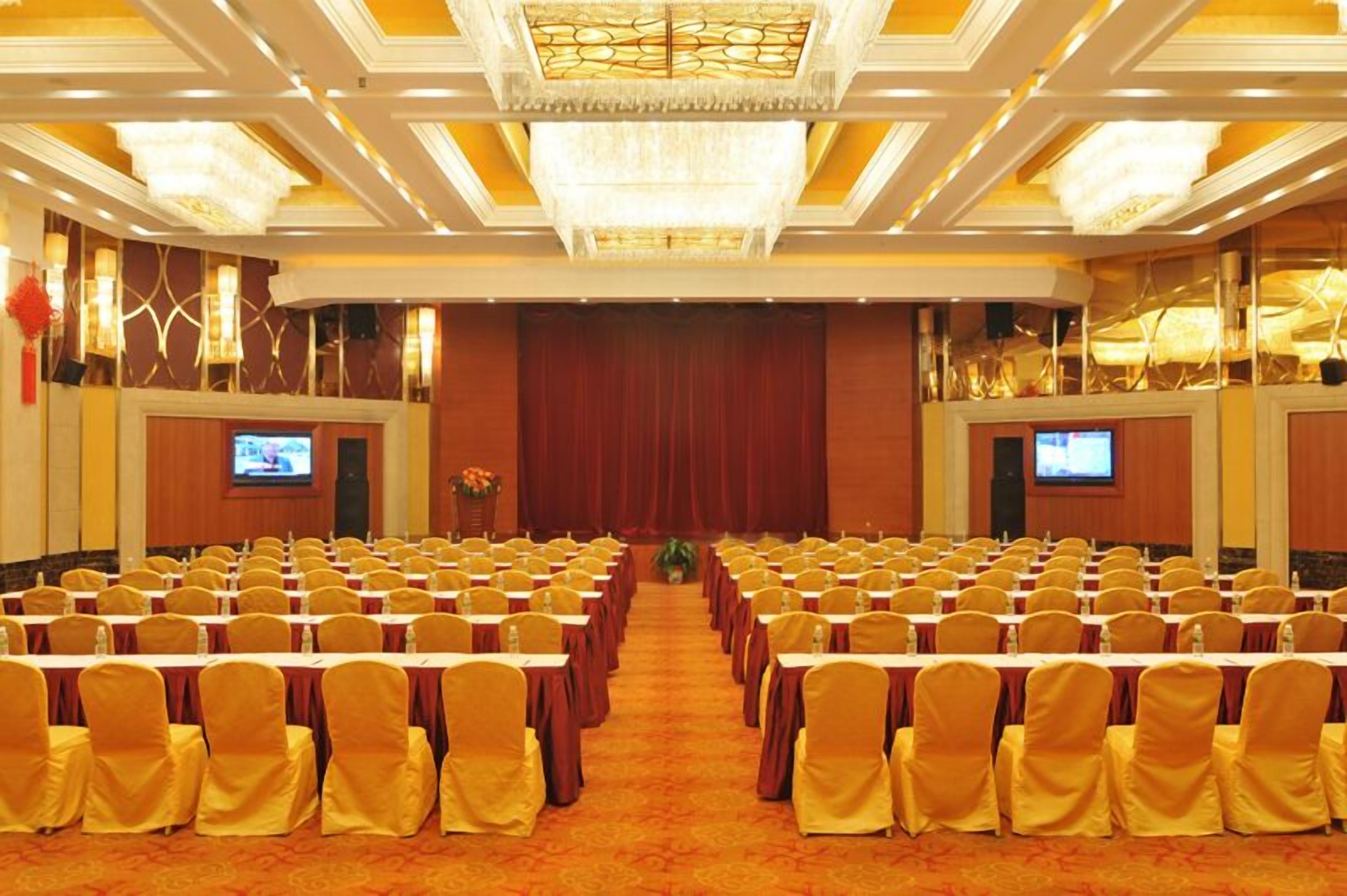 A conference room with rows of tables and chairs, a stage at the front, and two screens on either side.
