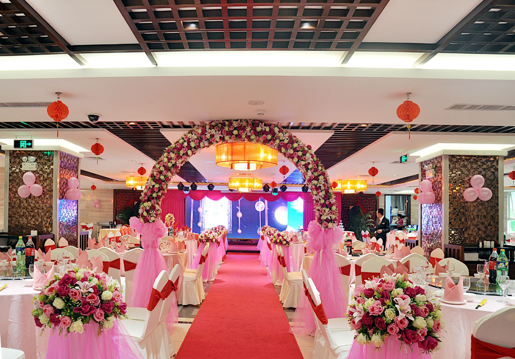 A banquet hall with a red carpet aisle, white chairs with pink sashes, and floral decorations.
