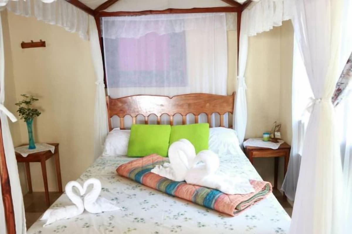 Ein ordentlich bezogenes Bett mit einer blumigen Tagesdecke, einem Holzkopfschild und einem grünen Kopfkissen.