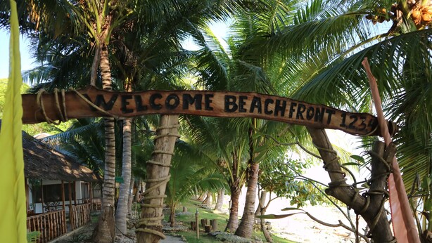 A wooden sign reading "WELCOME BEACHFRONT 1234" amidst a tropical setting with palm trees and a path leading to a building.