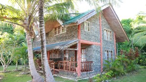 A two-story stone house with a thatched roof, surrounded by lush greenery and palm trees.