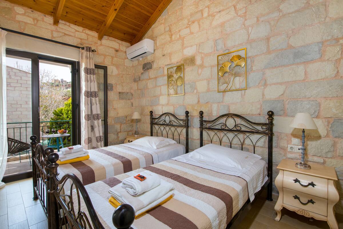 A room with two beds, a stone wall, a balcony with a table and chairs, and a window with curtains.