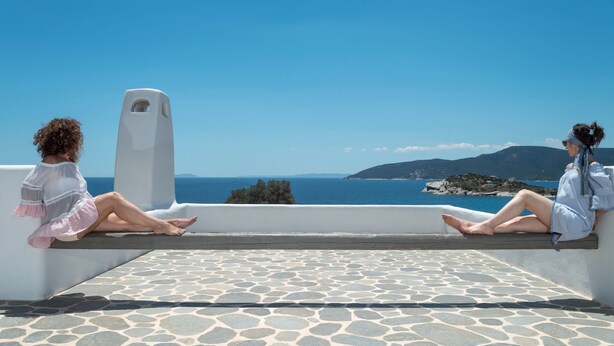 Deux personnes sont assises sur un balcon blanc dont le sol est en pierre, d’où elles peuvent admirer un paysage côtier avec des montagnes et la mer.