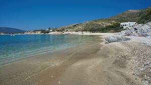 Une plage de galets aux eaux claires, une mer calme et un bâtiment blanc orné d’arcades, situé sur une colline.