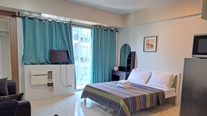 Quarto de hotel com cama, escrivaninha, cadeira, televisão e janela com cortinas.