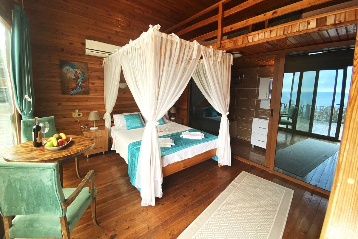 A wooden cabin bedroom with a four-poster bed, a dining table with fruit, and a view of the ocean through large sliding doors.