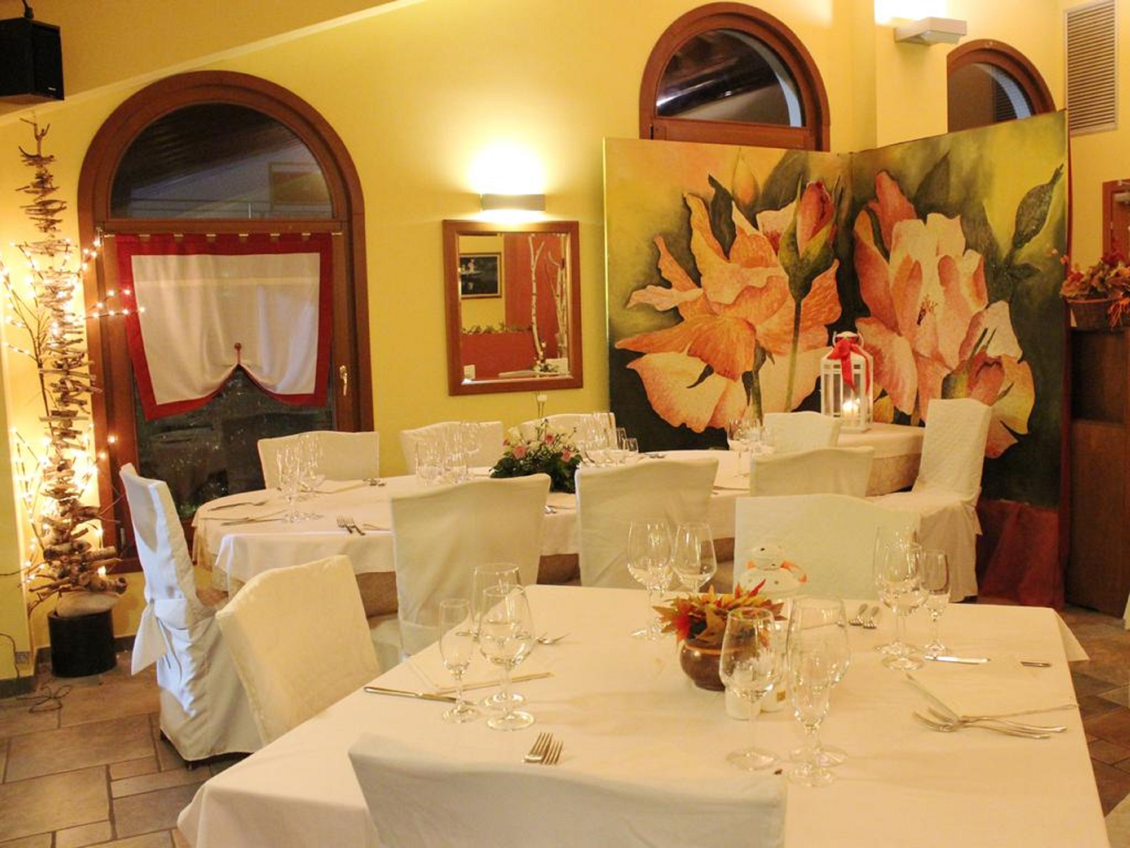 Un comedor con mesas dispuestas para una comida, con manteles blancos, sillas y un cuadro grande con motivos florales en la pared.