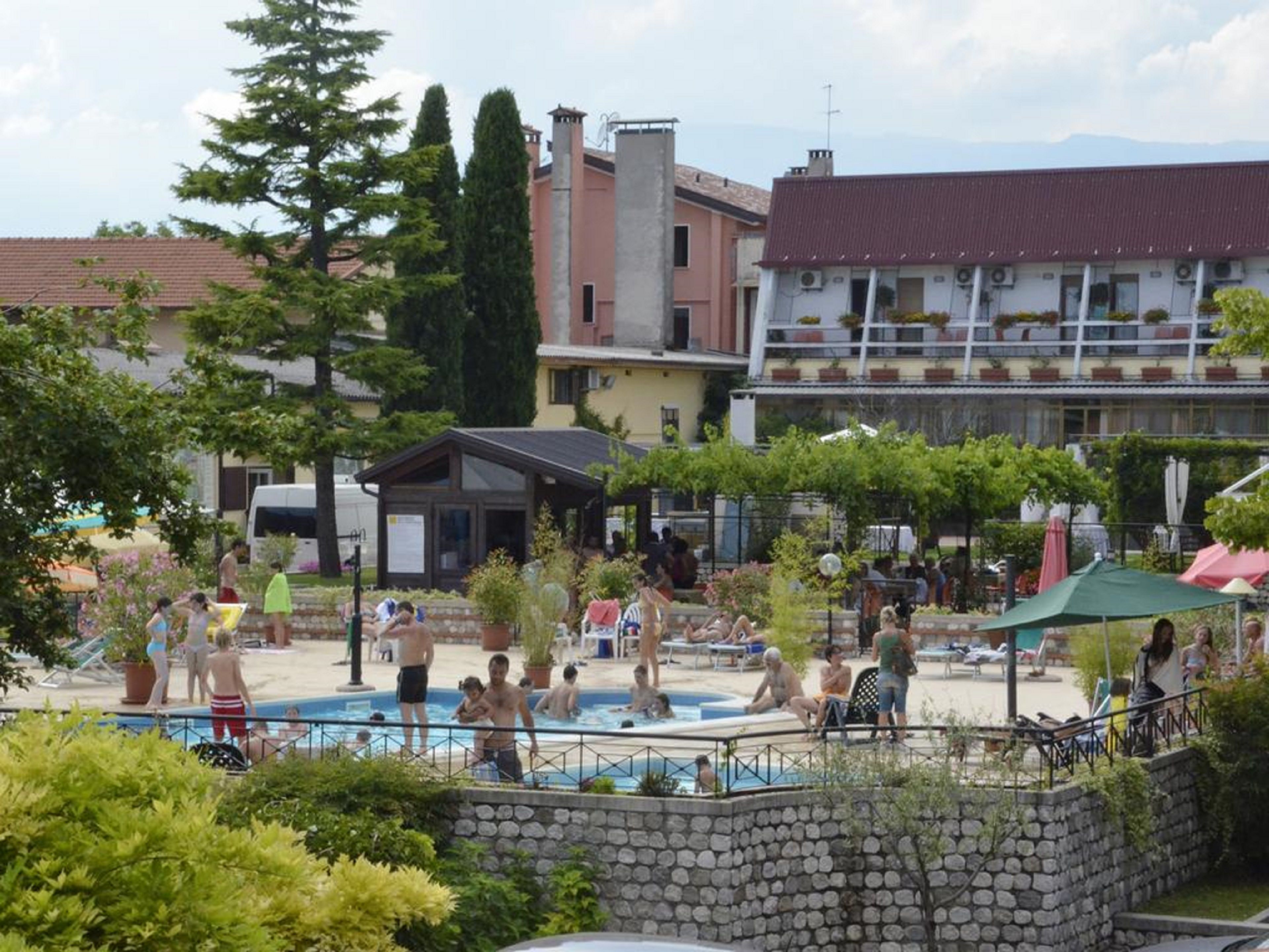 Zona de piscina con gente, rodeada de edificios y vegetación.