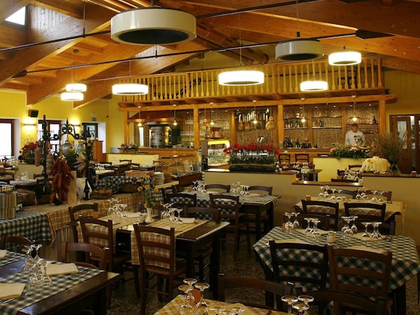 A restaurant with multiple dining tables, wooden beams on the ceiling, and a bar area with shelves of bottles.