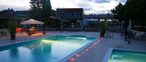 A swimming pool with a tiled deck, surrounded by lounge chairs and umbrellas, and a building in the background.