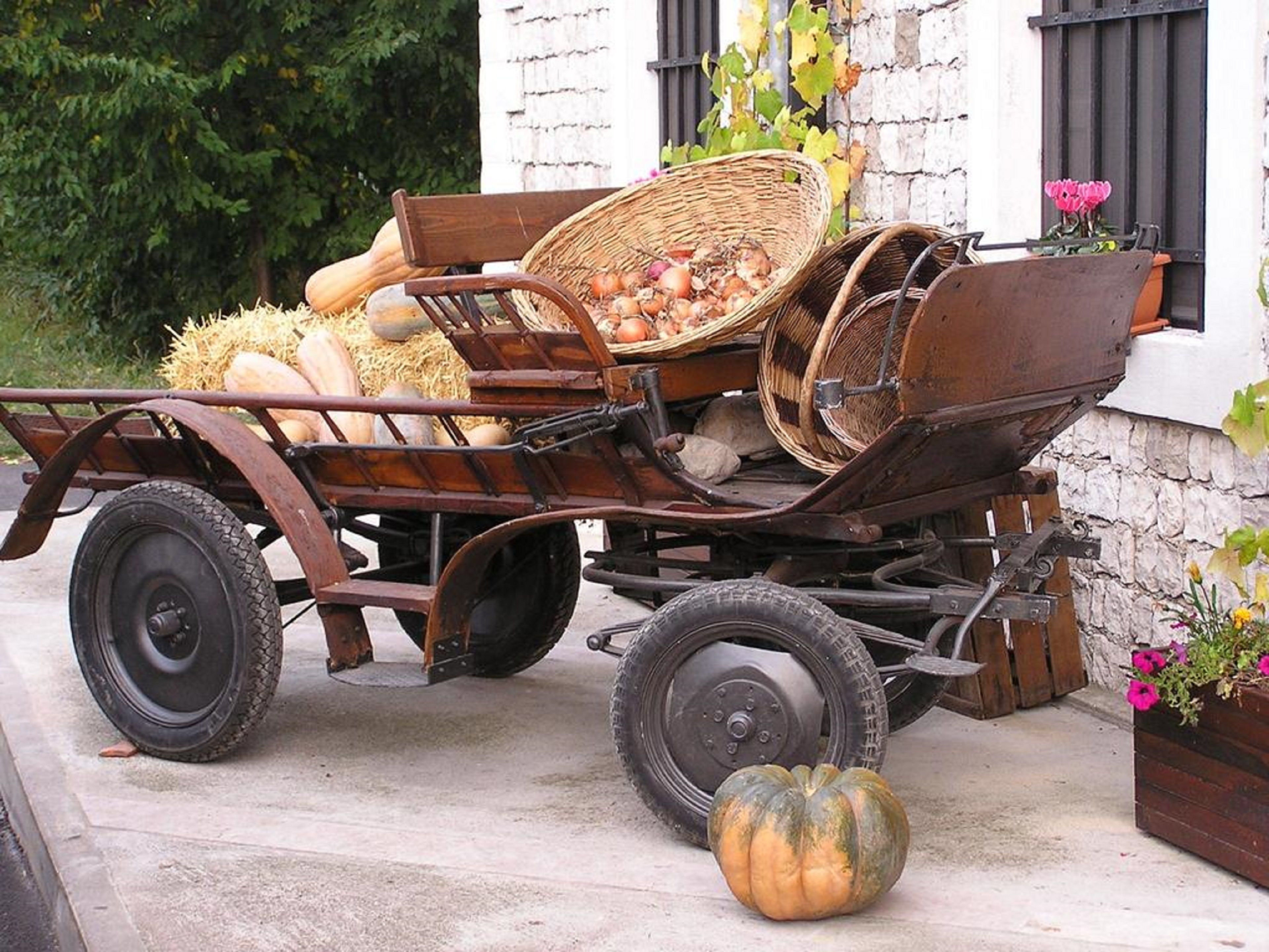 A vintage wooden cart with baskets and hay, parked in front of a white building with a stone wall and a window with black bars.