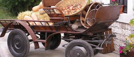 A vintage wooden cart with baskets and hay, parked in front of a white building with a stone wall and a window with black bars.