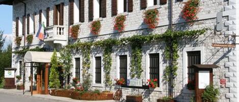 Il s’agit d’un bâtiment en pierre doté de nombreuses fenêtres, dont certaines sont pourvues de volets, et d’un balcon. Des jardinières remplies de fleurs rouges ornent les rebords des fenêtres.
