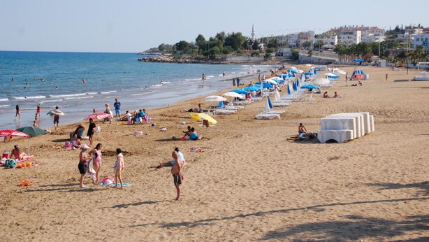 A beach with people sunbathing, swimming, and walking along the shore. There are umbrellas and lounge chairs lined up on the beach. - Hakan Apart Hotel (Erdemli)