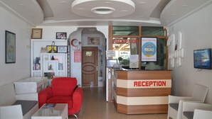 A reception area with a reception desk, a red armchair, and white chairs. There are framed pictures on the wall and a clock. - Hakan Apart Hotel (Erdemli)