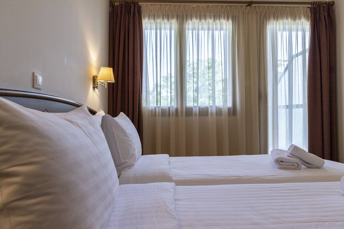 Una habitación de hotel con dos camas, una mesita de noche con lámpara y una ventana con cortinas.