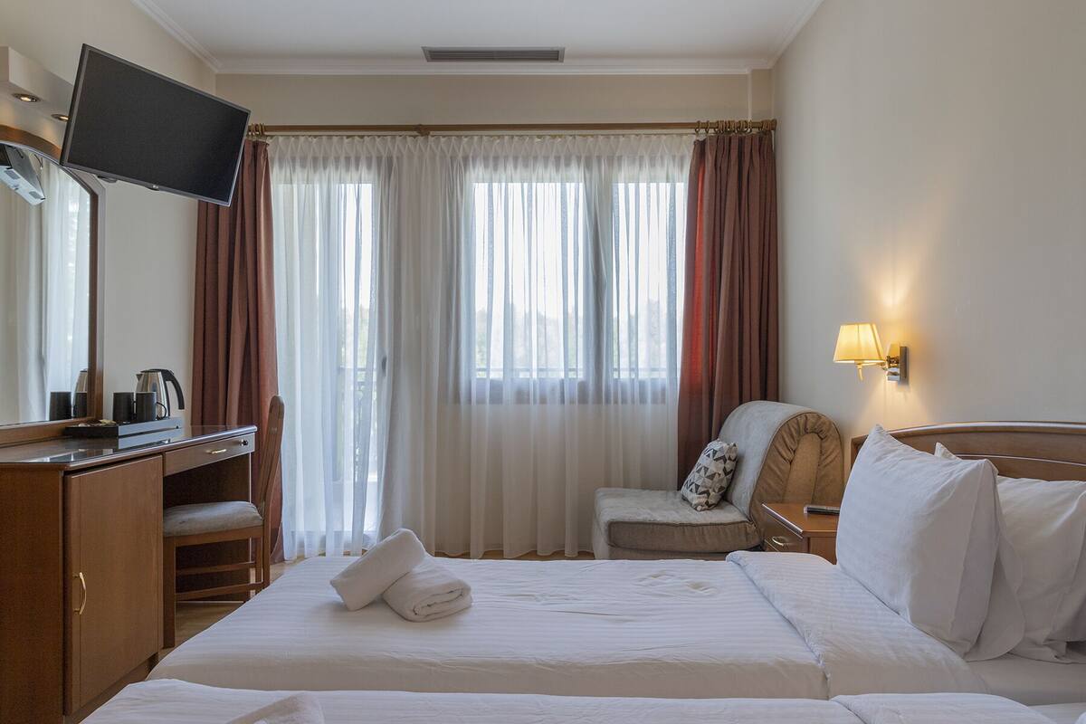 Una habitación de hotel con una cama, un escritorio con televisor, una silla y una mesita de noche con lámpara.