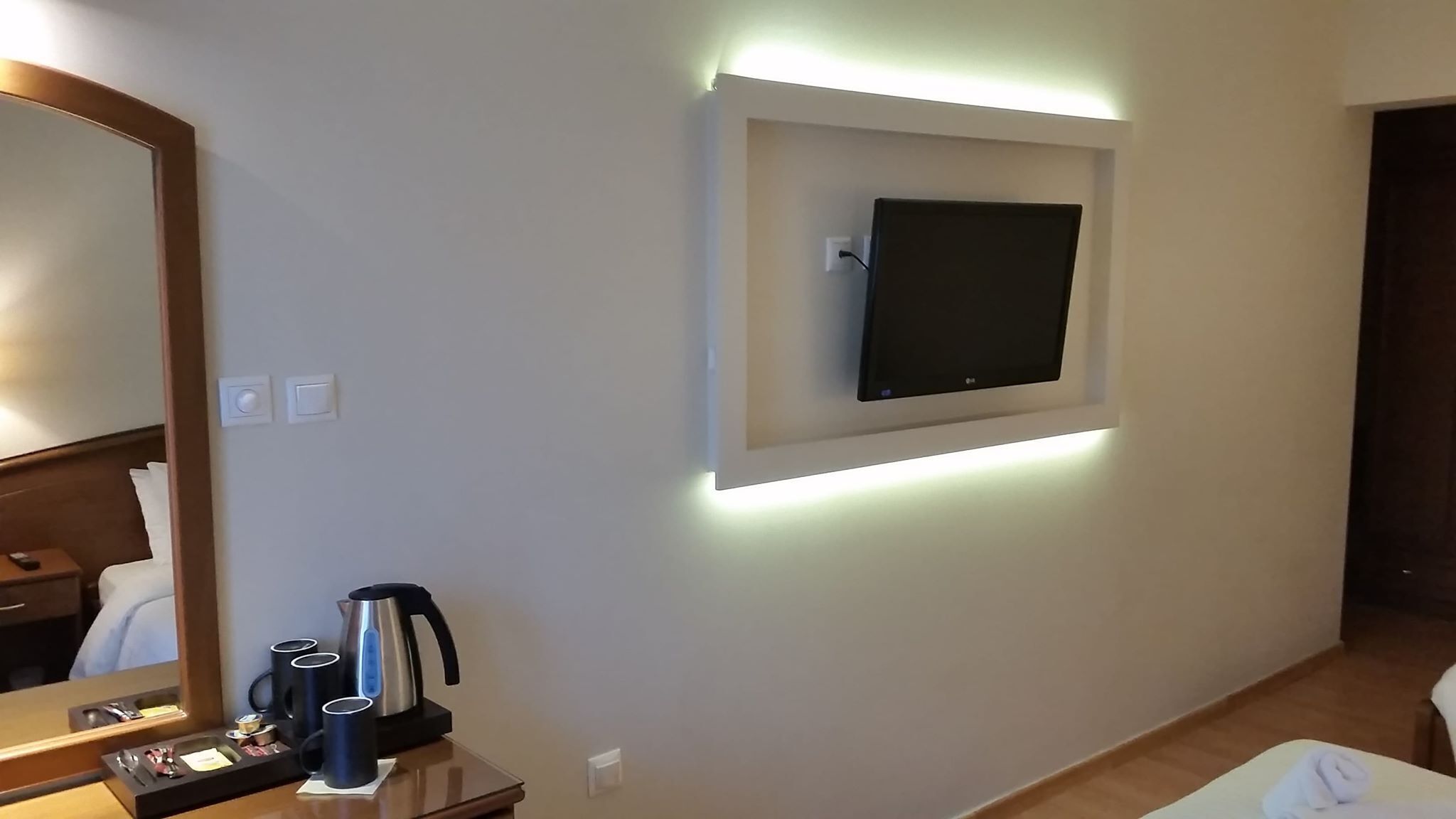 Une chambre d’hôtel dotée d’un téléviseur à écran plat fixé au mur, d’une table de chevet avec une bouilloire, des tasses et un plateau contenant des articles de toilette.