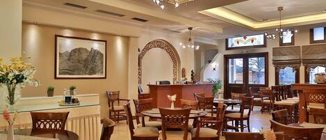A hotel lobby with multiple dining tables and chairs, a reception desk, and a chandelier.