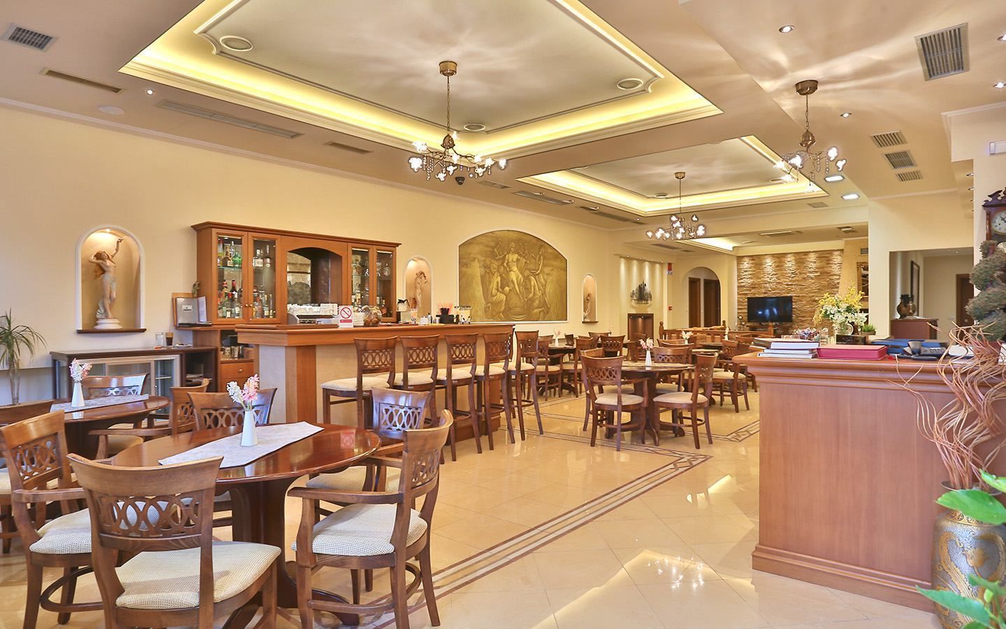 A spacious hotel lobby with multiple dining tables and chairs, a reception desk, and a bar area.