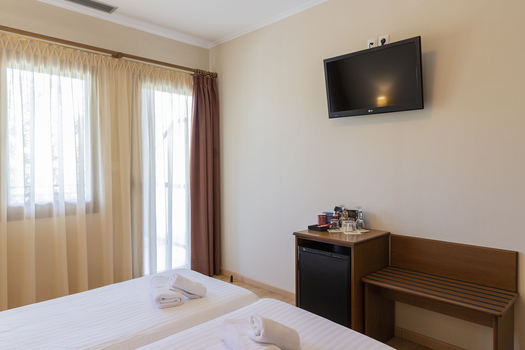 A hotel room with a bed, a TV mounted on the wall, a small desk with a coffee machine, and a wooden bench.