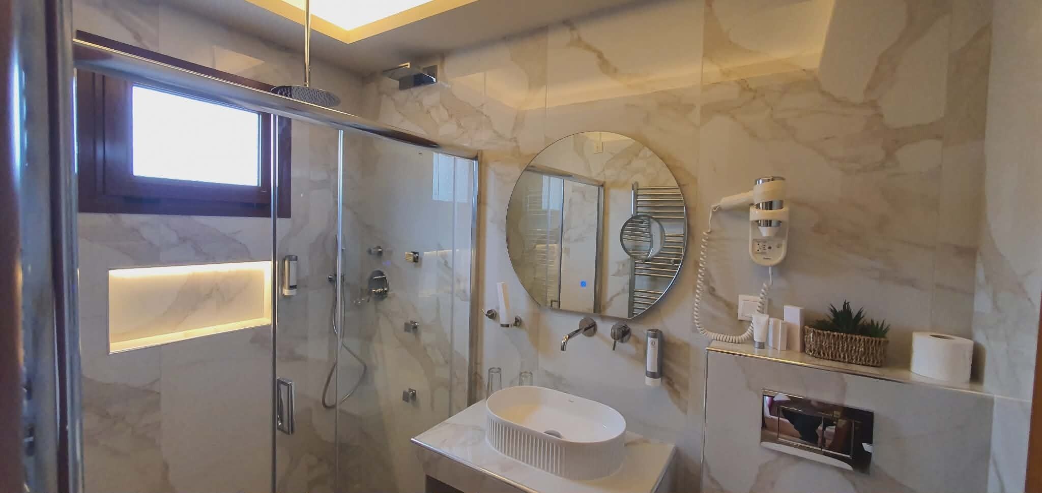 Un baño moderno con ducha envuelta en vidrio, un espejo redondo, un lavamanos blanco y paredes de mármol.