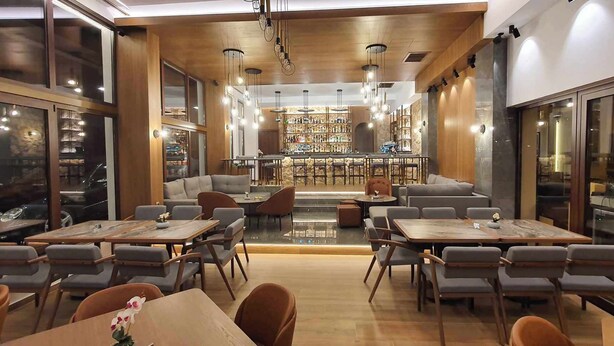 A modern restaurant with multiple dining tables and chairs, a bar area, and a view of the city through large windows. - Xenios Dias Boutique Hotel (Dio-Olympos)