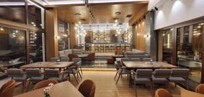 A modern restaurant with multiple dining tables and chairs, a bar area, and a view of the city through large windows.
