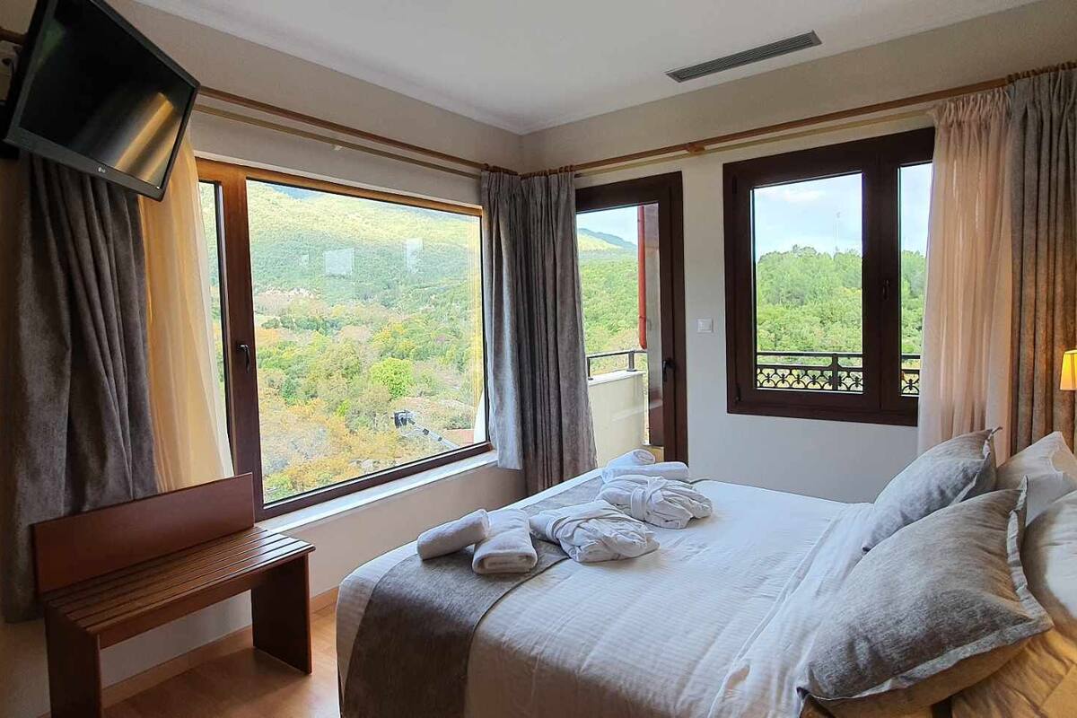 Habitación de hotel con cama, televisor montado en la pared y grandes ventanales con vistas al exterior.