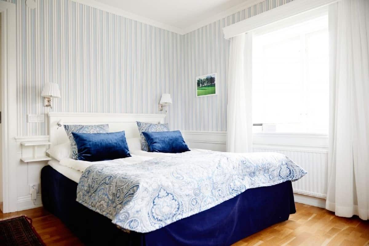 A neatly made bed with a blue and white patterned bedspread, a white headboard, and two blue pillows. There is a small picture on the wall, a window with white curtains, and a wooden floor.