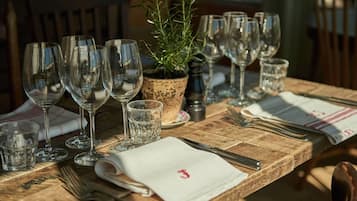 Una mesa de madera dispuesta con copas de vino, vasos de agua y cubiertos, con una planta en maceta en el centro.