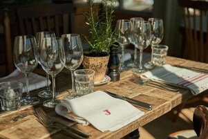 Una mesa de madera dispuesta con copas de vino, vasos de agua y cubiertos, con una planta en maceta en el centro.