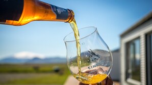 Un vaso transparente con un logotipo siendo llenado con un líquido dorado, al aire libre, con un edificio y montañas al fondo.