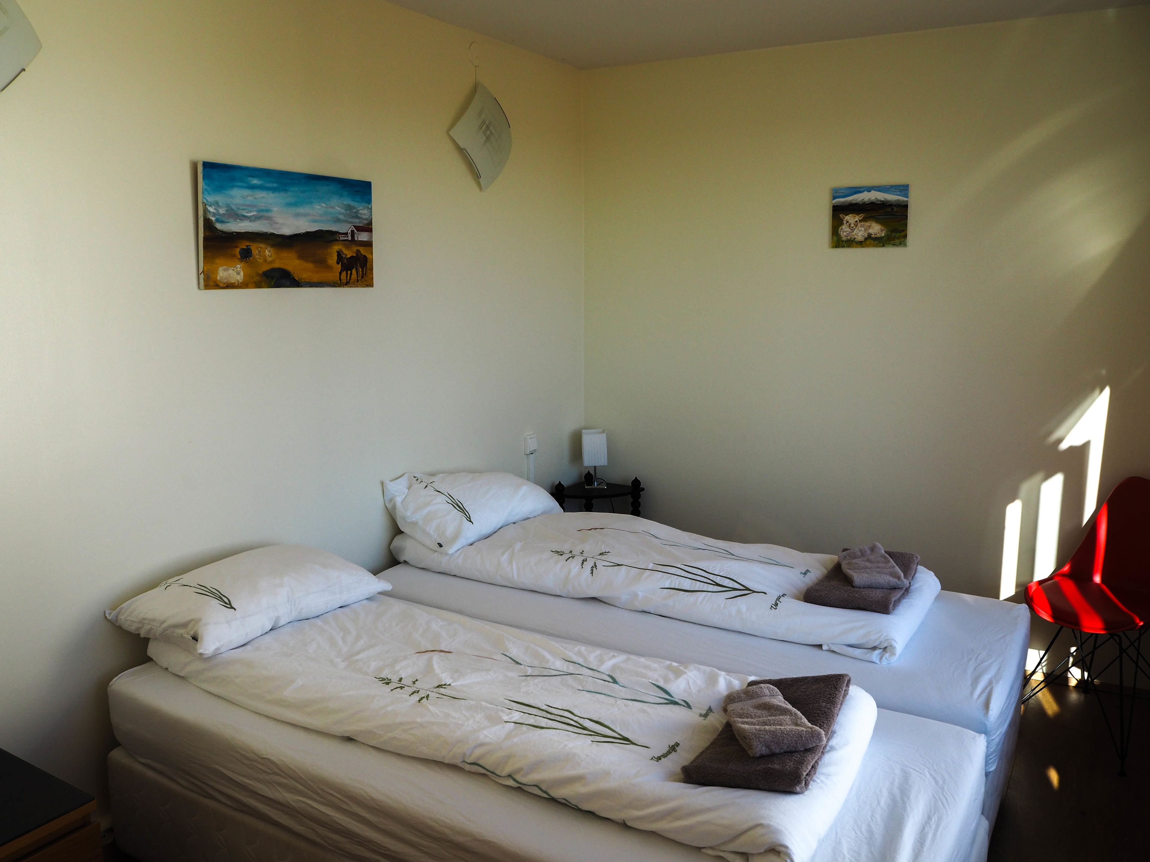 Une chambre avec deux lits, chacun agrémenté de draps blancs et de coussins décoratifs, une petite table de chevet avec une lampe, et deux tableaux accrochés au mur.