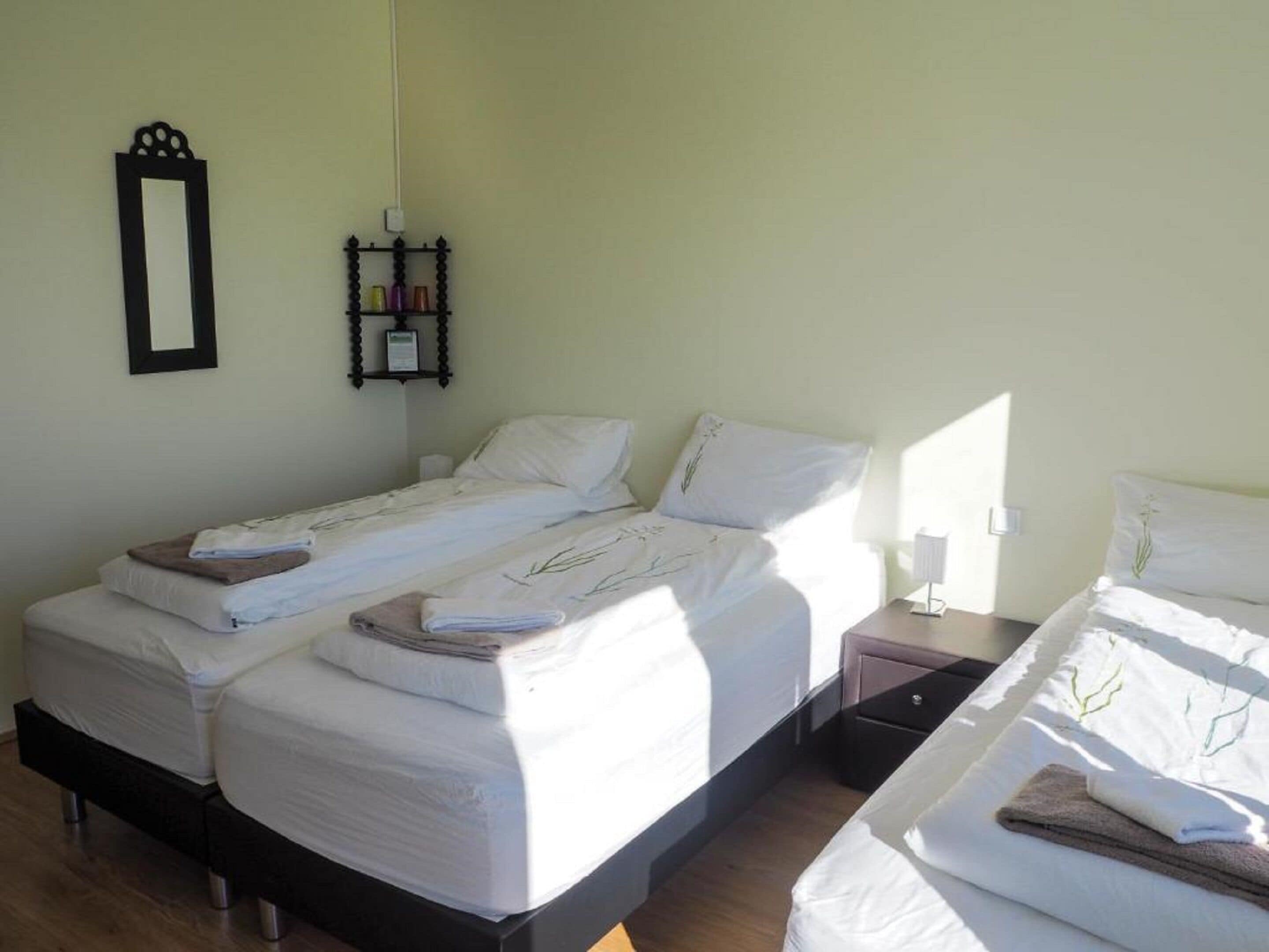 A hotel room with two single beds, a bedside table with a lamp, a small shelf with toiletries, and a wall-mounted mirror.