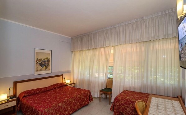 A hotel room with two beds, a chair, a desk with a lamp, and a painting on the wall.