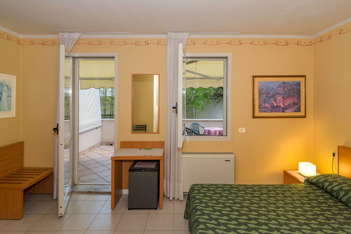 Hotelkamer met een bed, een bureau, een stoel, een tafeltje, een spiegel, een raam met uitzicht op groen en een schilderij aan de muur.