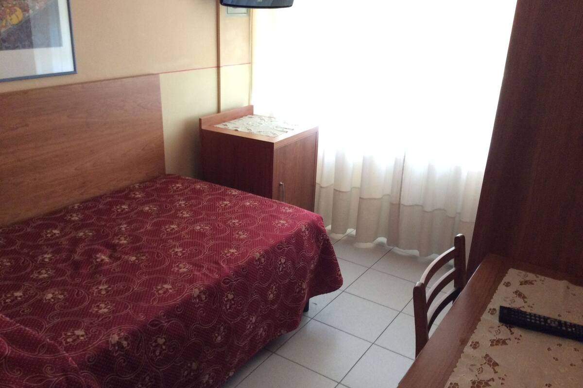 Een kleine hotelkamer met een enkel bed, een houten nachtkastje, een aan de muur bevestigde televisie en een raam met gordijnen.