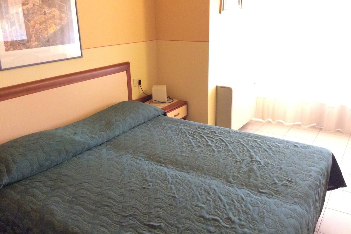 Een slaapkamer met een bed, nachtkastje, lamp en een aan de muur hangend schilderij.