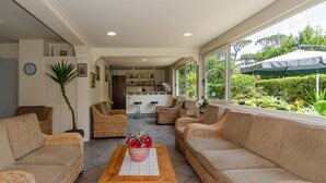 Amplia sala de estar con muebles de mimbre, una mesa de centro con un centro de mesa floral y vistas a un jardín a través de grandes ventanales.