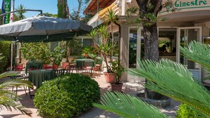 Un patio al aire libre con mesas y sillas, rodeado de plantas en macetas y árboles.