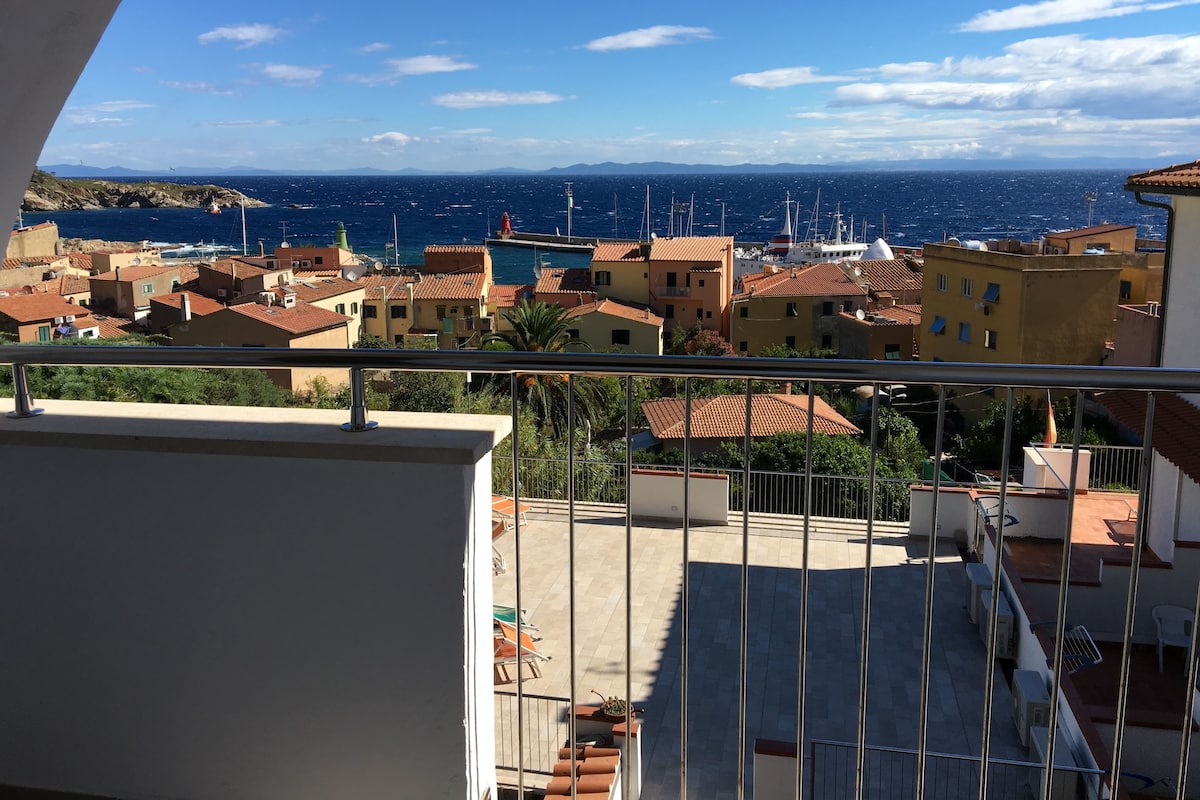 A balcony with a view of a coastal town, featuring terracotta-roofed buildings and a clear blue sky.