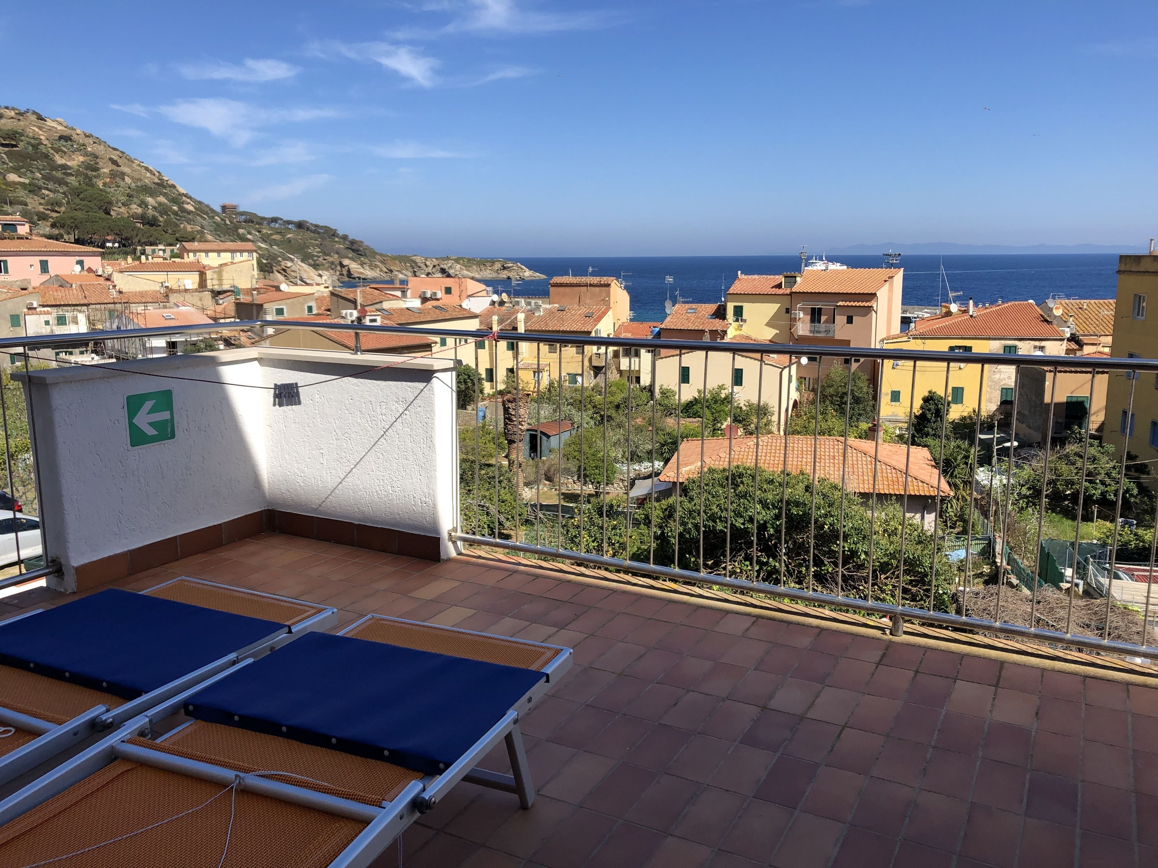 A terrace with a view of a coastal town, including buildings with red-tiled roofs and a clear blue sky.