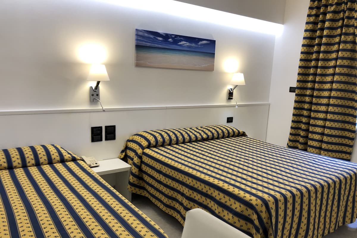 A hotel room with two beds, each with a striped bedspread, a nightstand, a lamp, a painting of a beach scene, and a curtain with a pattern.