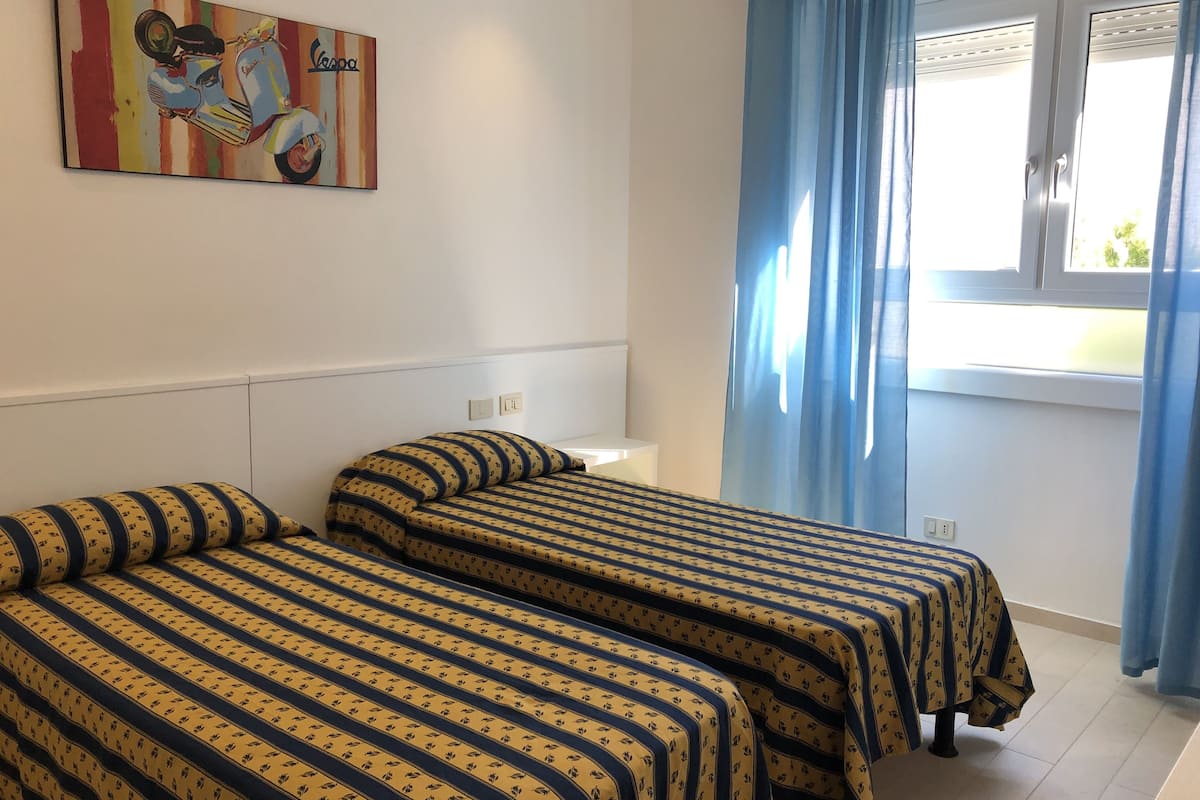 A hotel room with two beds, a window with blue curtains, and a painting on the wall.