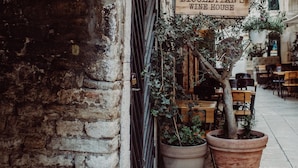 Una calle estrecha con un edificio que dice "DIOCLECIAN'S WINE HOUSE". Se ven macetas con plantas y mesas al aire libre.