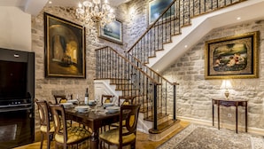 Un comedor con una mesa y sillas de madera, una araña y una escalera con un muro de piedra de fondo.